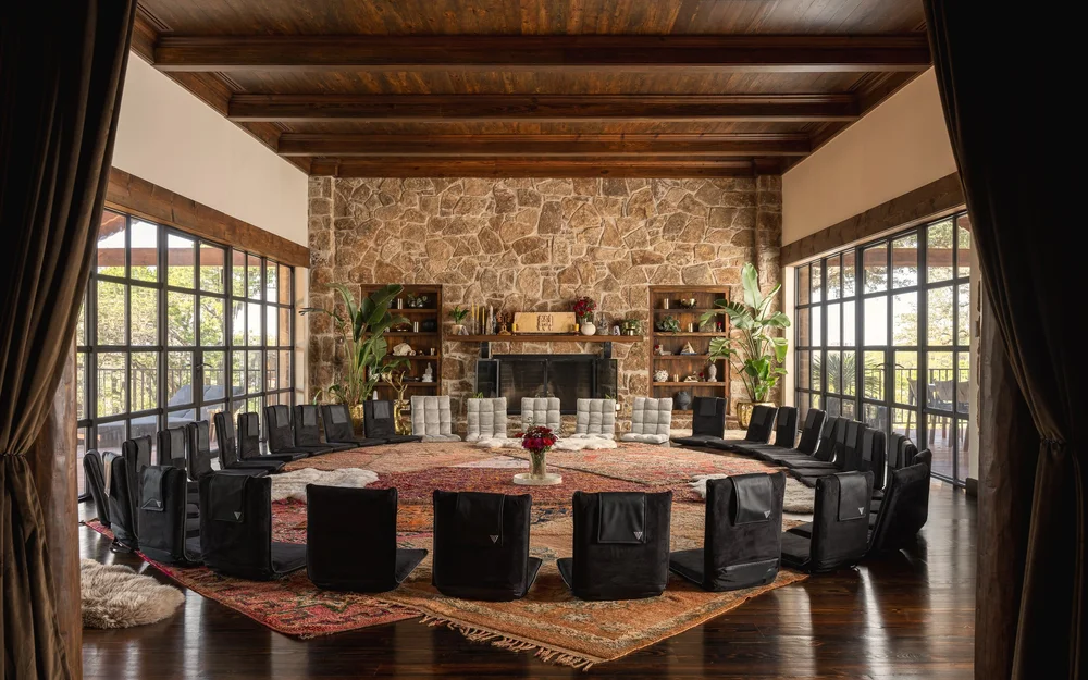The TICo ceremony room: a circle of reclining chairs arranged on a patterned rug before a stone fireplace, floor-to-ceiling windows opening onto the Texas Hill Country