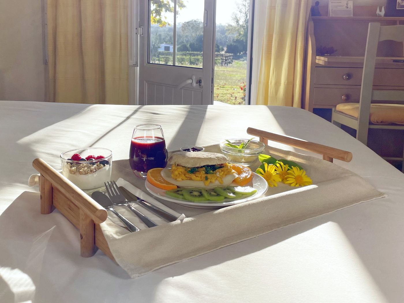 Breakfast tray on a bed in a Tabula Rasa Portugal guest room — yogurt with berries and granola, juice, scrambled eggs on toasted English muffin, sliced kiwi and orange, with morning light streaming through an open door to the grounds